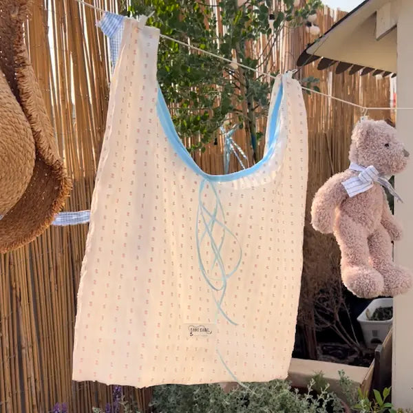 A large handmade fabric tote bag in cream with a small dot print and blue binding, hanging on an outdoor clothesline beside a straw hat and a teddy bear