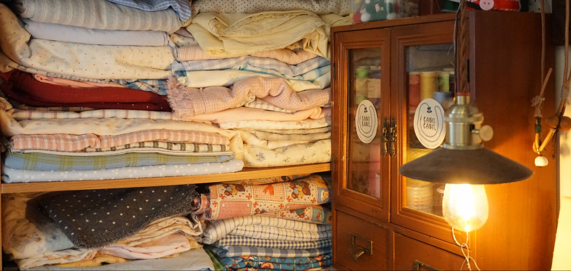 Shelves of neatly folded handmade fabric textiles in soft pastel colors, with a warm-lit wooden cabinet and Kang Kang Handmade labels