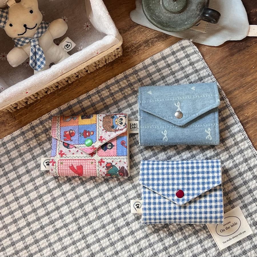 Three Kang Kang handmade Cottage Card Cases on a plaid mat — Colorful Patchwork with green snap on the left, Blue Denim Bow Embroidery with silver snap on the upper right, and Blue Gingham with red snap on the lower right, with a bear plush toy in the background