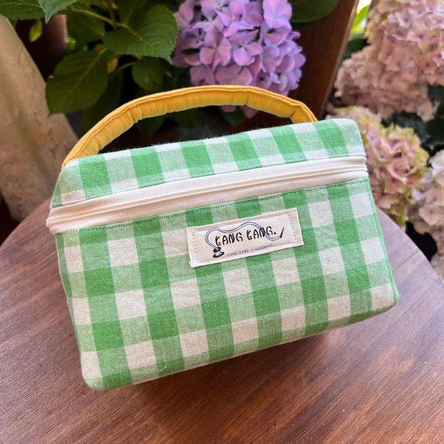 Kang Kang handmade Colorblock Wash Bag in green and white gingham fabric with yellow contrast handle, displayed on a wooden table with purple and pink hydrangeas in the background