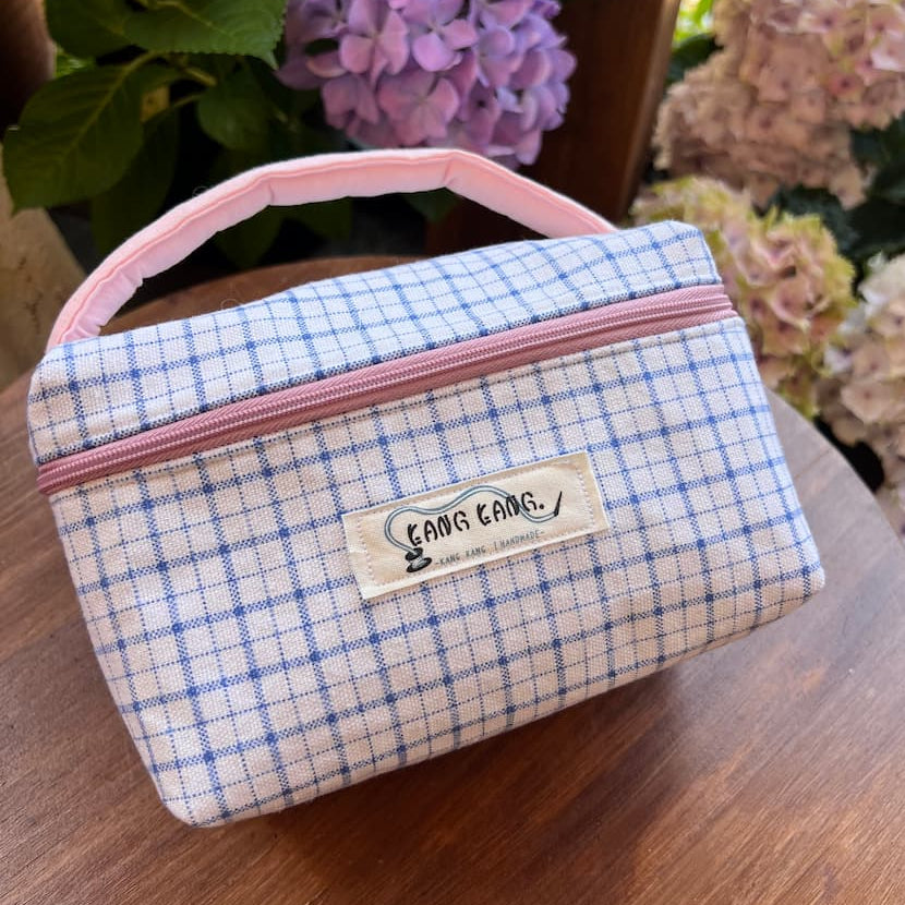 Kang Kang handmade Colorblock Wash Bag in blue and white fine check fabric with pink contrast handle, displayed on a wooden table with purple hydrangeas in the background