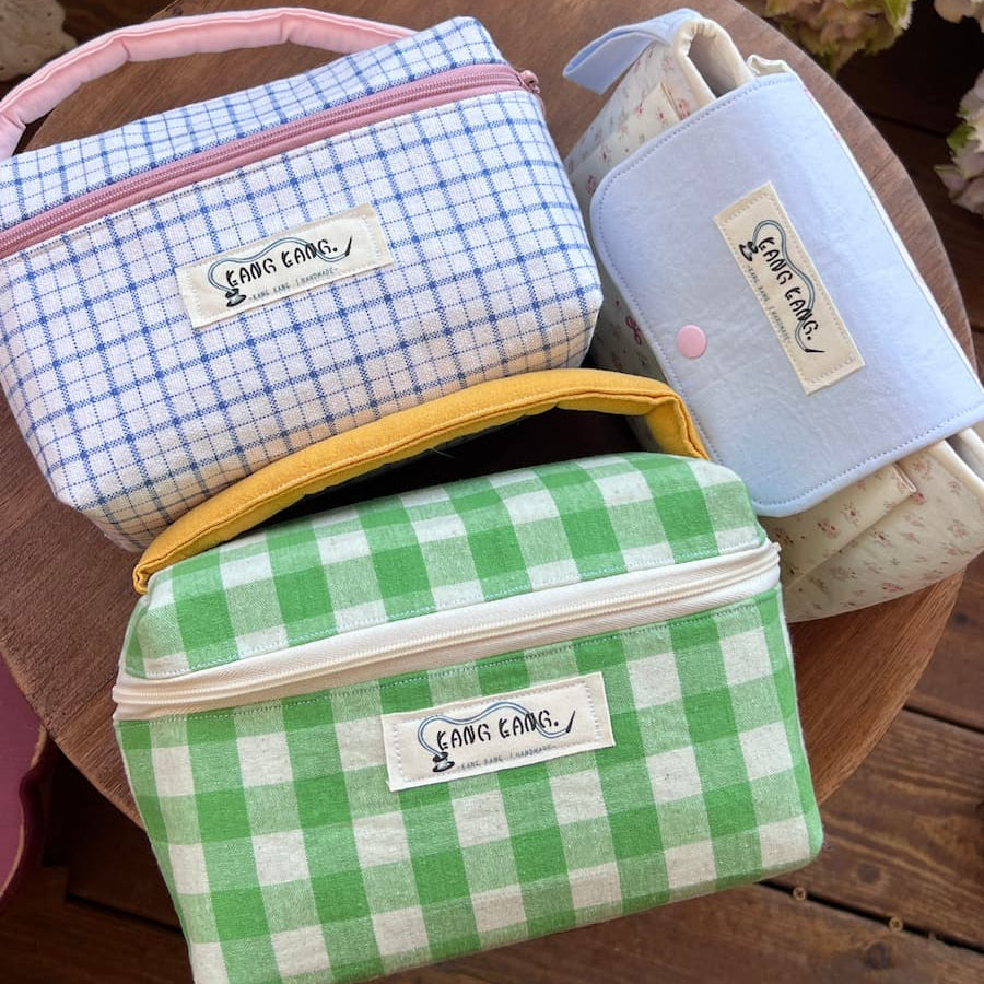 Three Kang Kang handmade Colorblock Wash Bags on a wooden tray — Blue Gingham with pink handle on the upper left, Green Gingham with yellow handle in the front, and a Flap Pouch in the upper right, with hydrangeas in the background