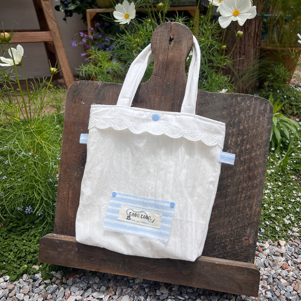 Kang Kang handmade Dreamy Lace Tote Bag in cream white fabric with scalloped lace trim and blue stripe front pocket, displayed on a wooden board in a garden with white cosmos flowers