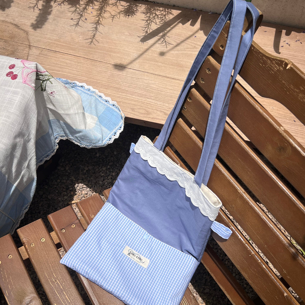 Kang Kang handmade Lace Trim Tote Bag in blue colorblock fabric with white lace trim, leaning against a wooden bench outdoors in sunlight