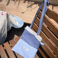 Kang Kang handmade Lace Trim Tote Bag in blue colorblock fabric with white lace trim, leaning against a wooden bench outdoors in sunlight