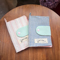 Two Kang Kang handmade Garden Plaid Passport Covers on a wooden table — Pink Stripe with mint green clasp on the left, Blue Plaid with mint green clasp on the right, with purple hydrangeas in the background