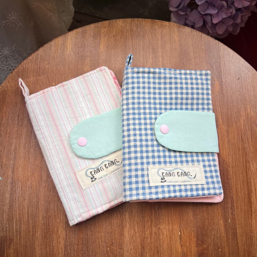 Two Kang Kang handmade Garden Plaid Passport Covers on a wooden table — Pink Stripe with mint green clasp on the left, Blue Plaid with mint green clasp on the right, with purple hydrangeas in the background
