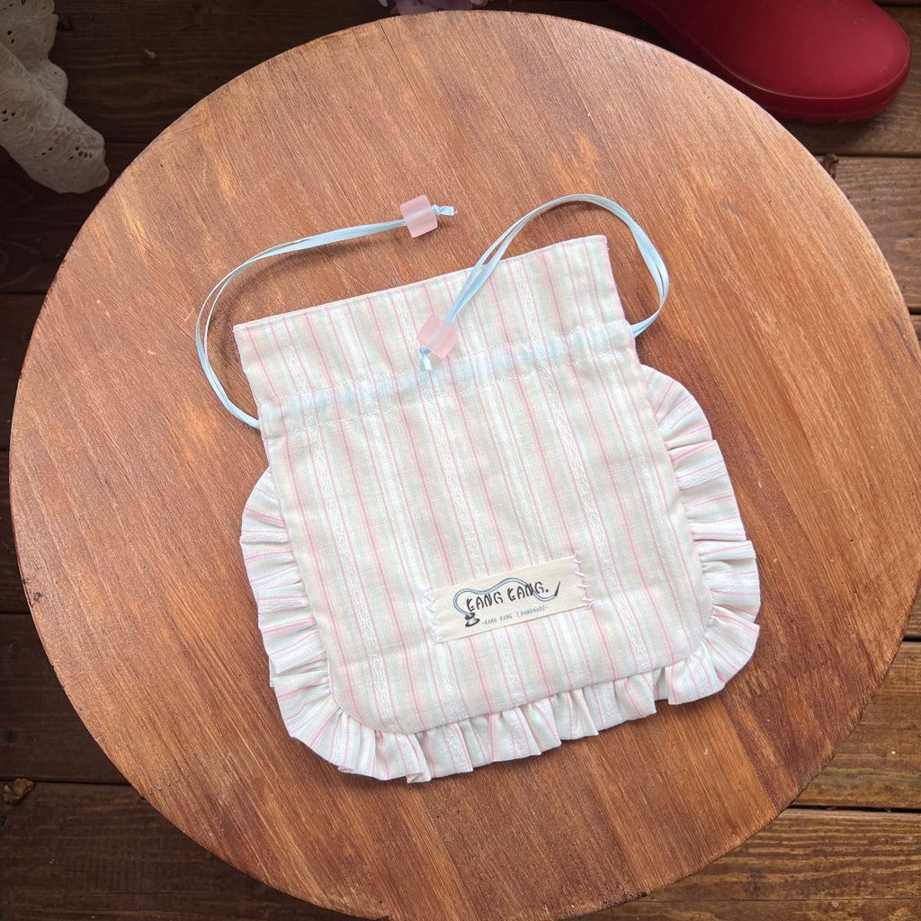 Kang Kang handmade Sugar Cube drawstring bag in pink stripe fabric with ruffled edges and mint blue ribbon, flat lay on wooden surface