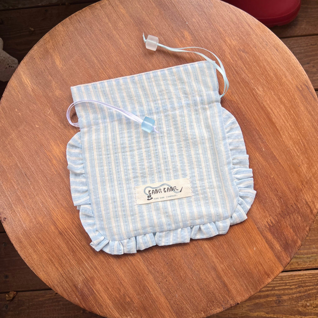 Kang Kang handmade Sugar Cube drawstring bag in light blue stripe fabric with ruffled edges and white ribbon, flat lay on wooden surface
