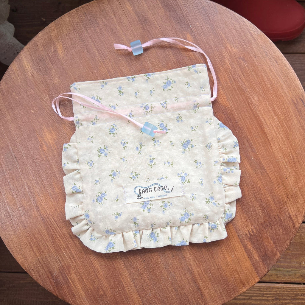 Kang Kang handmade Sugar Cube drawstring bag in butter yellow floral print with ruffled edges and pink ribbon, flat lay on wooden surface