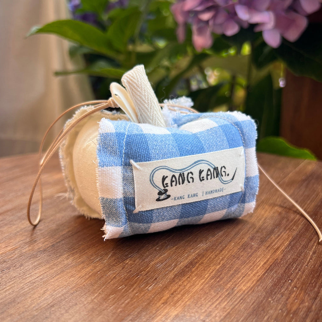 Kang Kang handmade Lucky Apple Charm in blue gingham fabric with cream stem and leather cord, displayed on a wooden surface with purple hydrangeas in the background