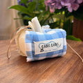 Kang Kang handmade Lucky Apple Charm in blue gingham fabric with cream stem and leather cord, displayed on a wooden surface with purple hydrangeas in the background