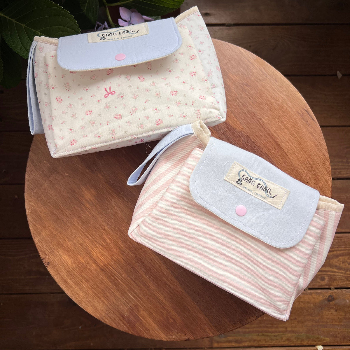 Two Kang Kang handmade Flap Pouch Bags on a round wooden table — Ditsy Floral in cream with blue flap and pink snap on top, Pink Stripe with white flap and pink snap in front