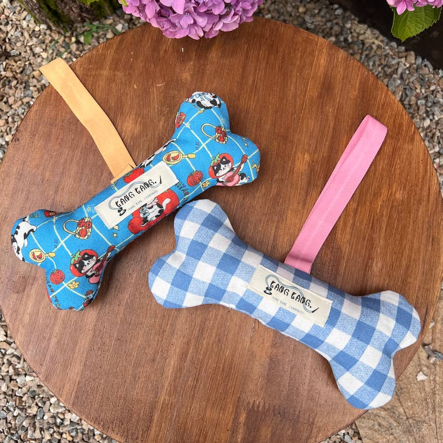 Two Kang Kang handmade bone-shaped dog toys on a wooden tray — Cat Print with elastic orange strap on the left, Gingham Check with pink strap on the right, with hydrangeas in the background