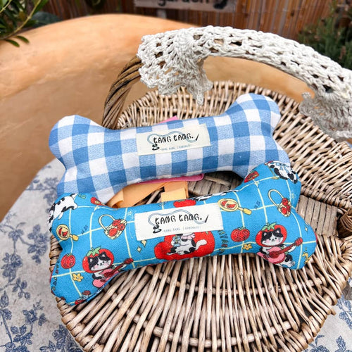Two Kang Kang handmade bone-shaped dog toys resting on a wicker basket — Gingham Check in blue and white on top, Cat Print in vibrant blue below, photographed outdoors
