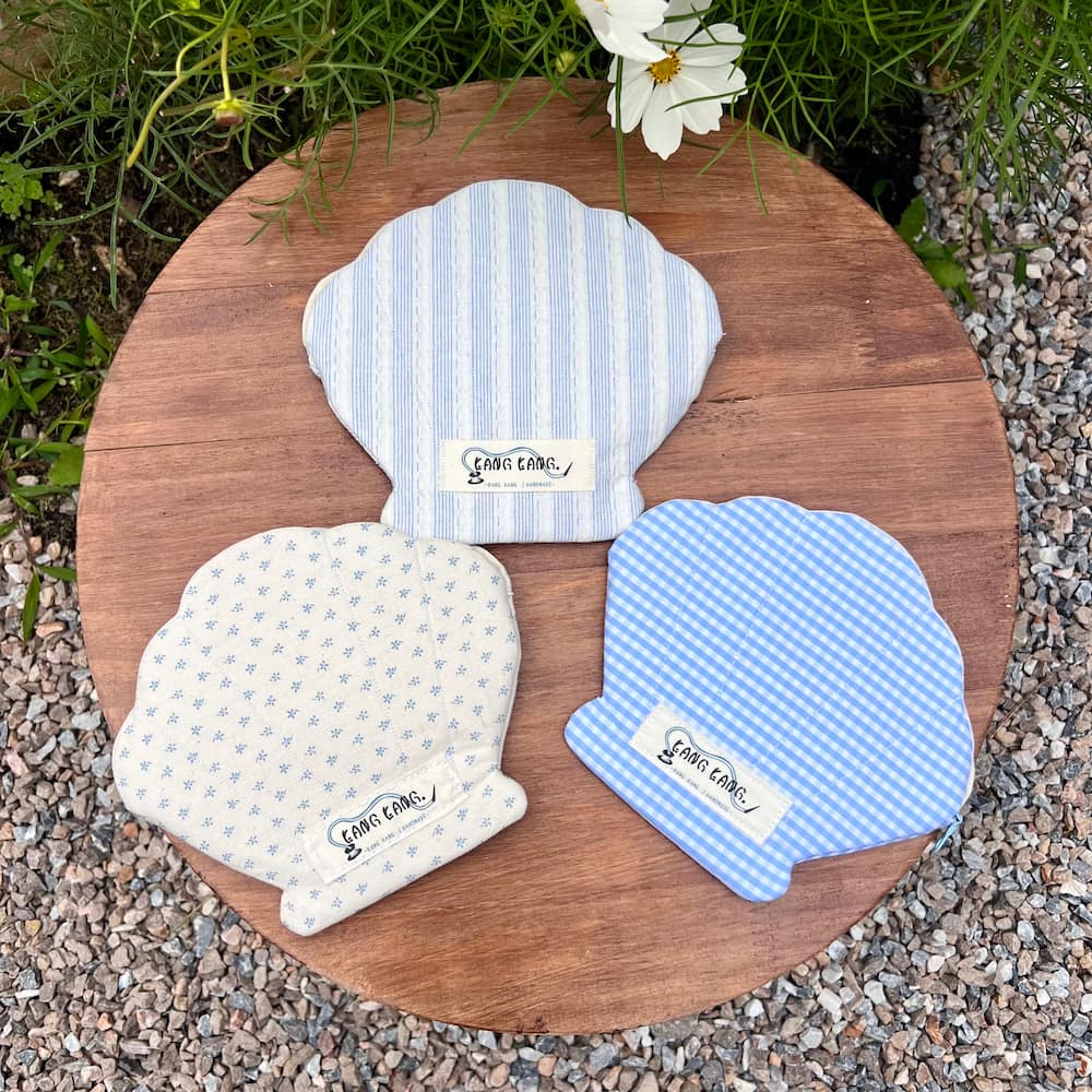 Three handmade shell-shaped fabric pouches by Kang Kang displayed on a round wooden board outdoors — blue stripe, cream floral, and blue gingham patterns