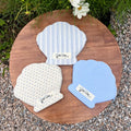 Three handmade shell-shaped fabric pouches by Kang Kang displayed on a round wooden board outdoors — blue stripe, cream floral, and blue gingham patterns
