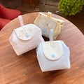 Three Kang Kang handmade Cloud Pouches on a round wooden table — Baby Pink with fairy tale flap on the left, Beige with Alice in Wonderland flap in the back, and Baby Blue with floral flap in the front, with green hydrangeas in the background