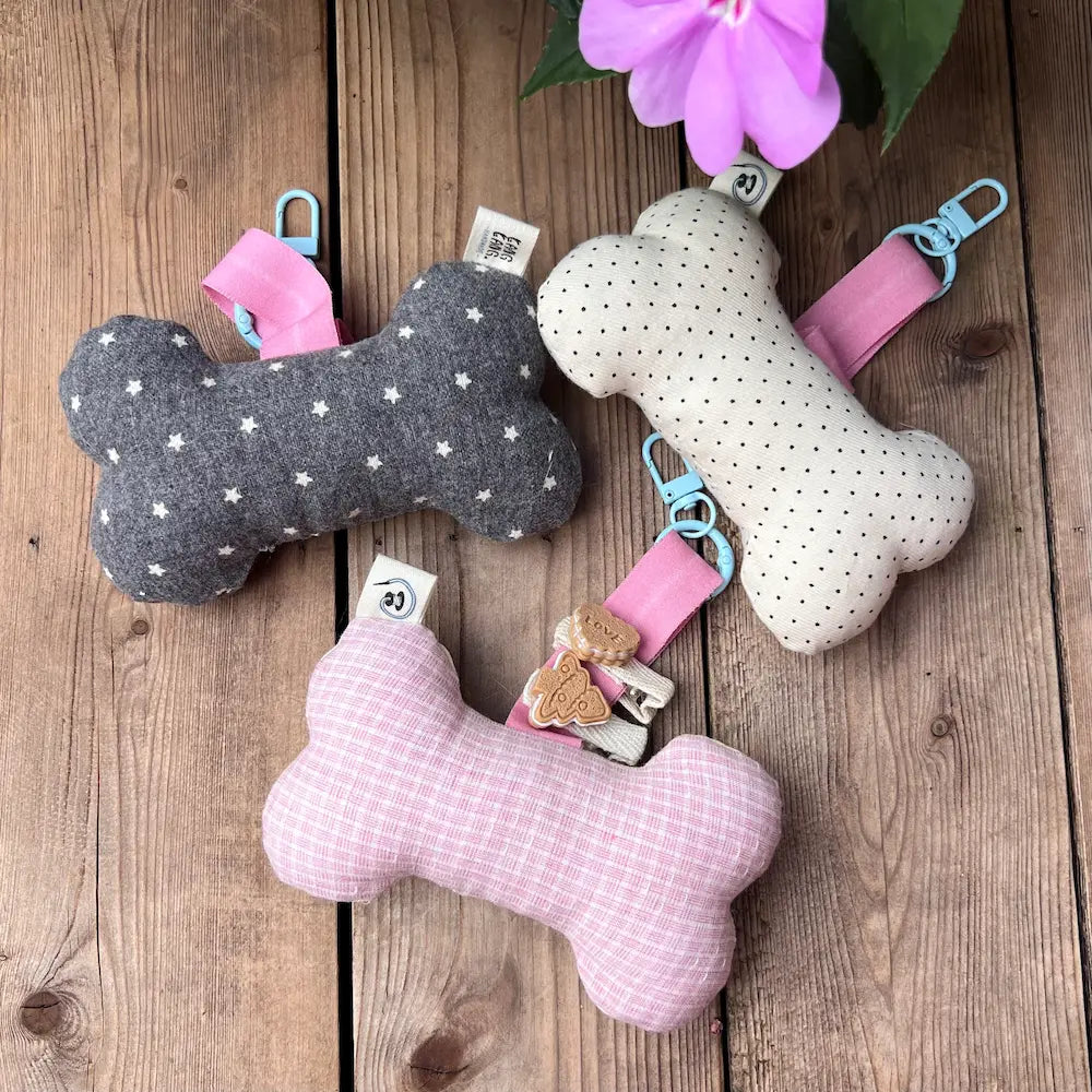 Three handmade bone-shaped dog toy keychains displayed on wooden surface with pink flowers — dark grey stars, cream polka dots, and light pink grid fabric styles