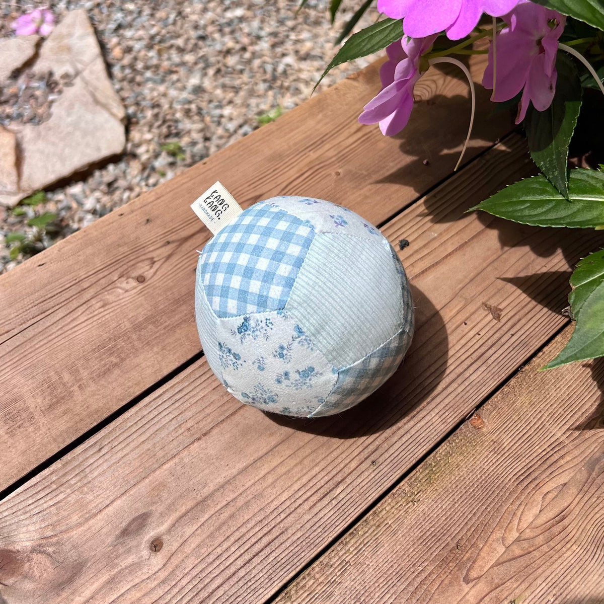 Kang Kang Handmade Patchwork Dog Ball on wooden deck outdoors with pink flowers in background, side view