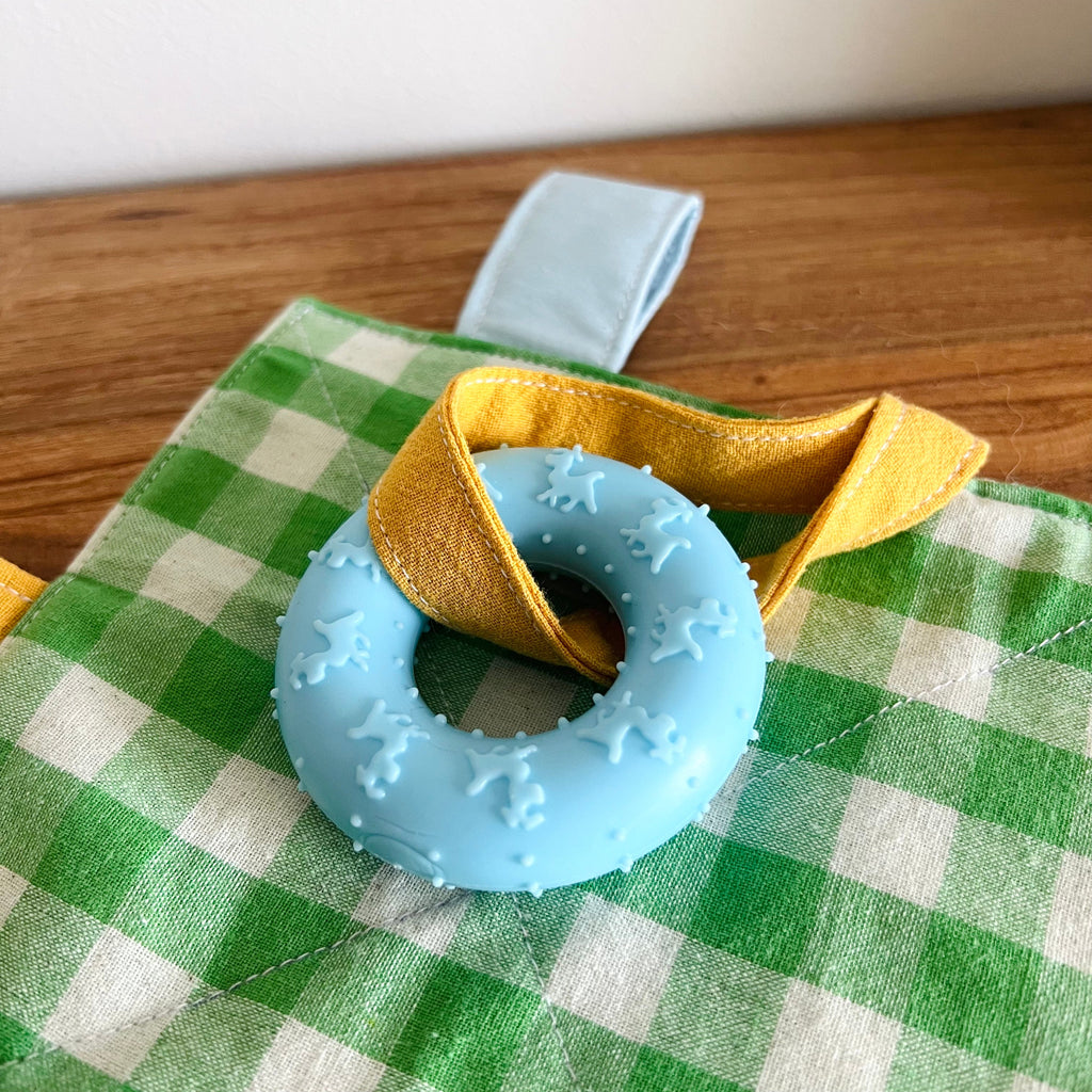 Close-up of blue silicone teething ring attached to the Crinkle Play Mat with yellow ribbon loop