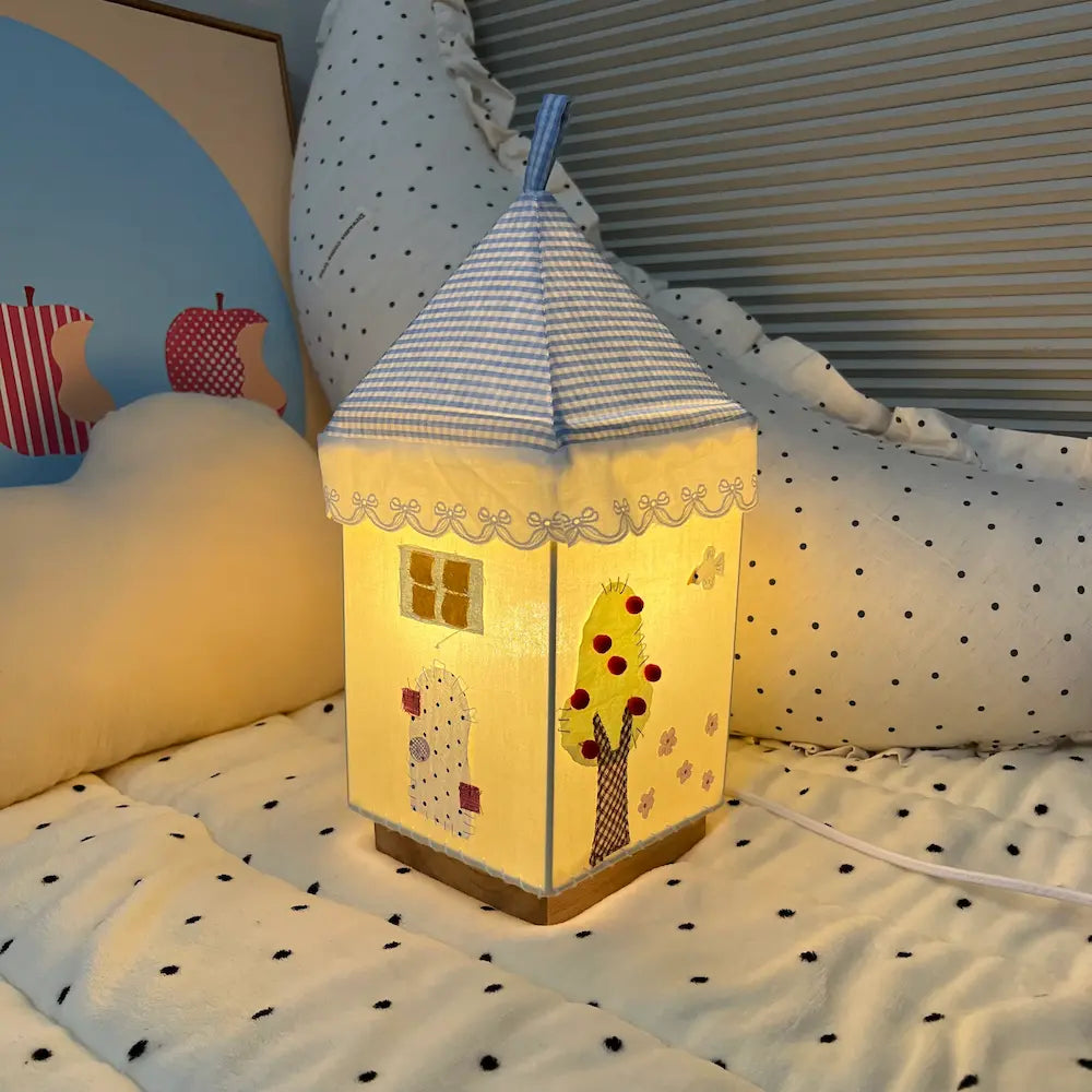Handmade fabric house lamp glowing with warm yellow LED light, showing garden scene with fruit tree and hedgehog appliqué, placed on a cozy bed