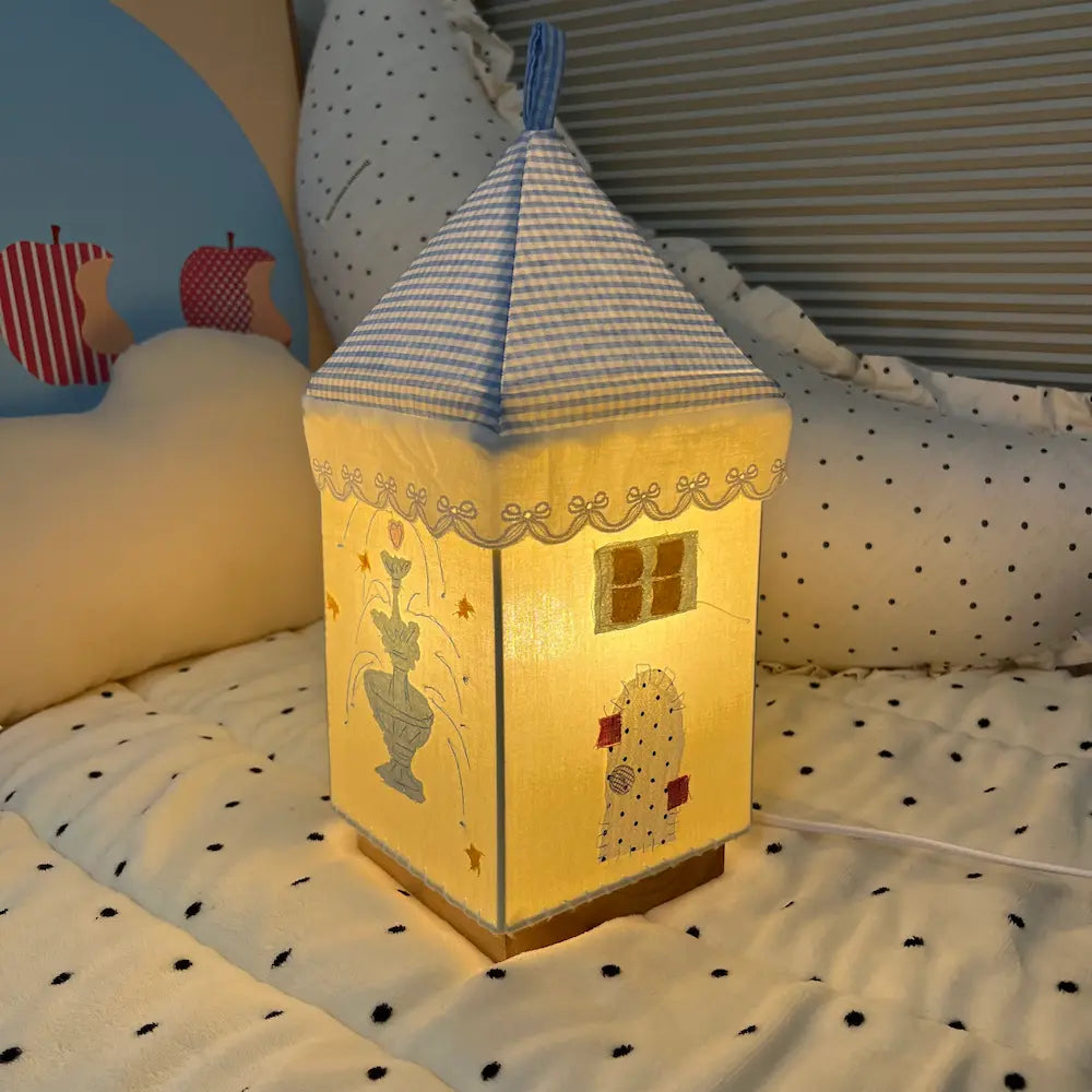 Handmade fabric house lamp lit up at night, displaying fountain scene with star details and a small figure, soft warm glow on bedside setting