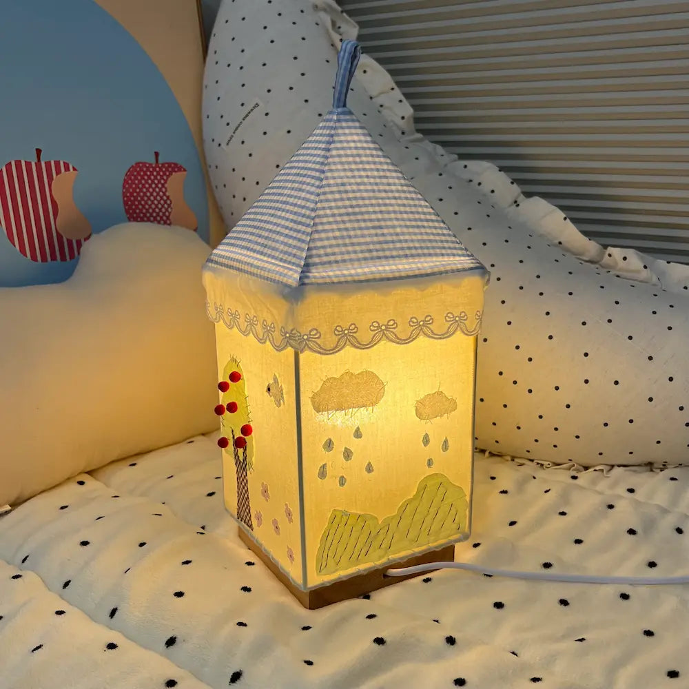Fabric house lamp showing rainy day scene with clouds and raindrops hand-stitched on the shade, warm amber light glowing through the fabric
